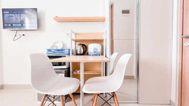 Une chambre d’hôtel de taille réduite, dotée d’un bureau en bois, de deux chaises blanches, d’une télévision et d’une kitchenette.