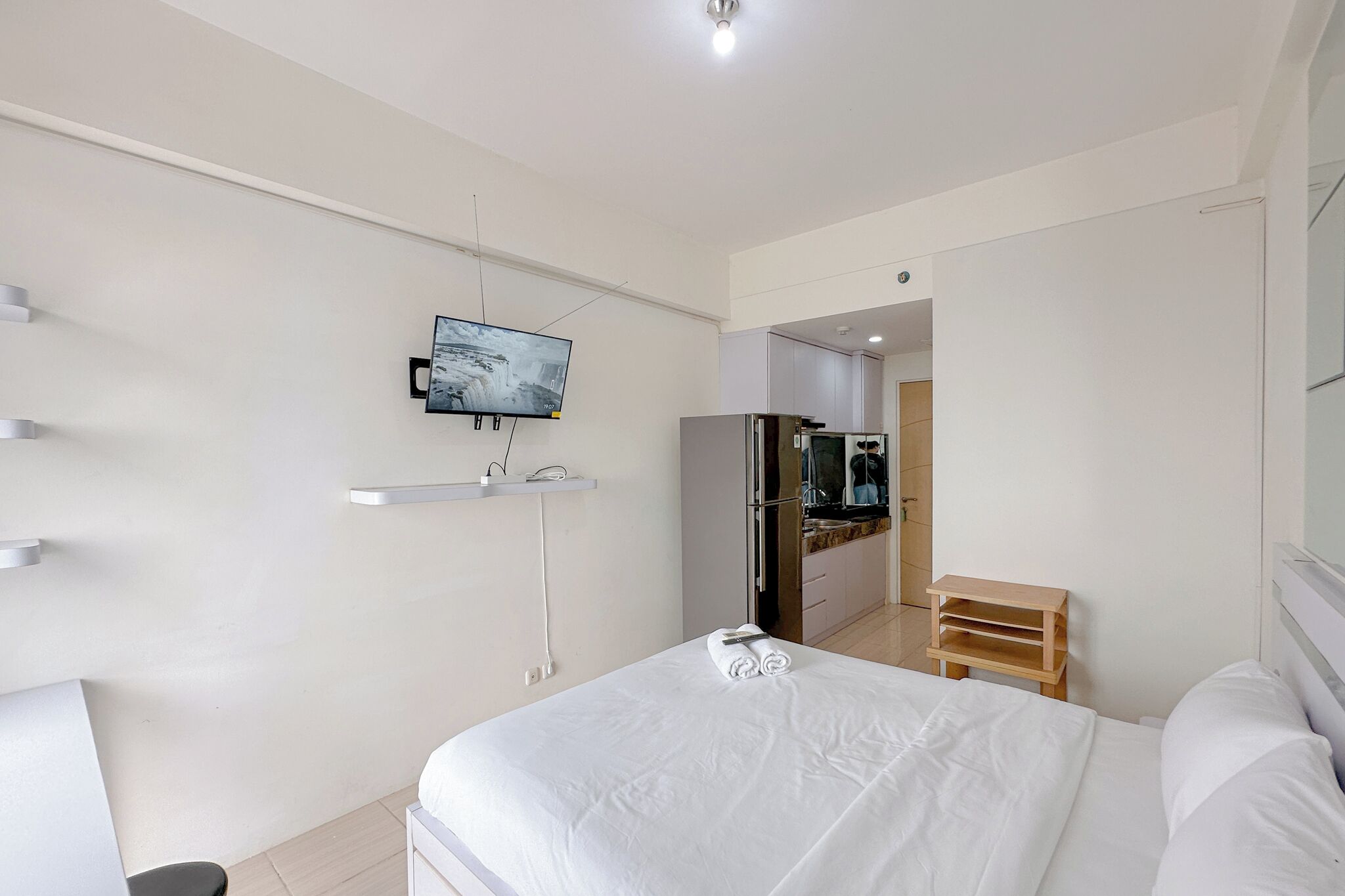 A modern hotel room with a large bed, a TV mounted on the wall, a small wooden side table, and a kitchenette in the background.