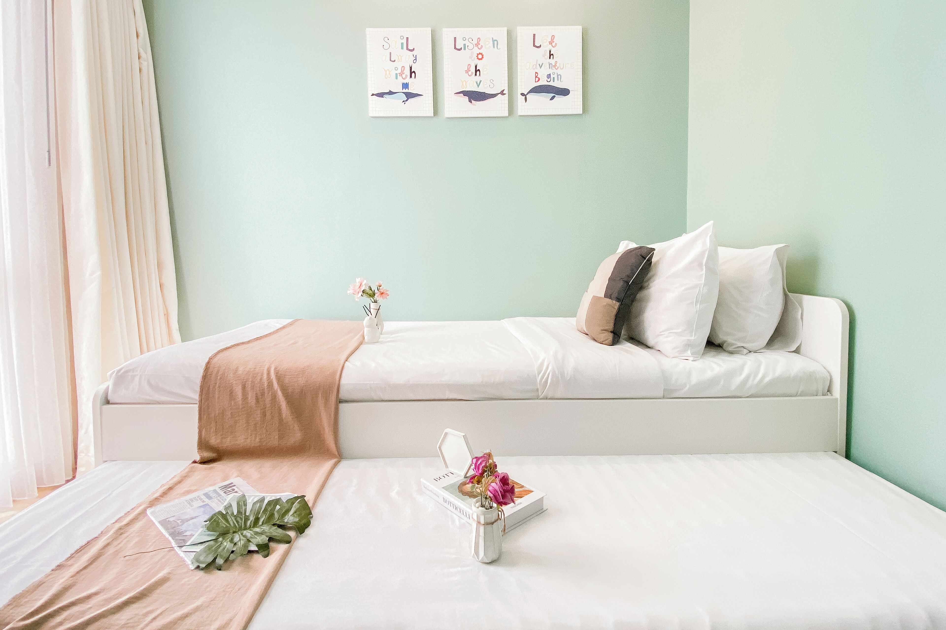 Un lit bien fait, recouvert d’une couette blanche et d’un plaid beige. Un petit vase blanc contenant des fleurs roses est posé sur le lit.