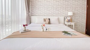 A hotel room with a white brick wall, a large bed with white bedding, a nightstand with a lamp, and a window with sheer curtains.