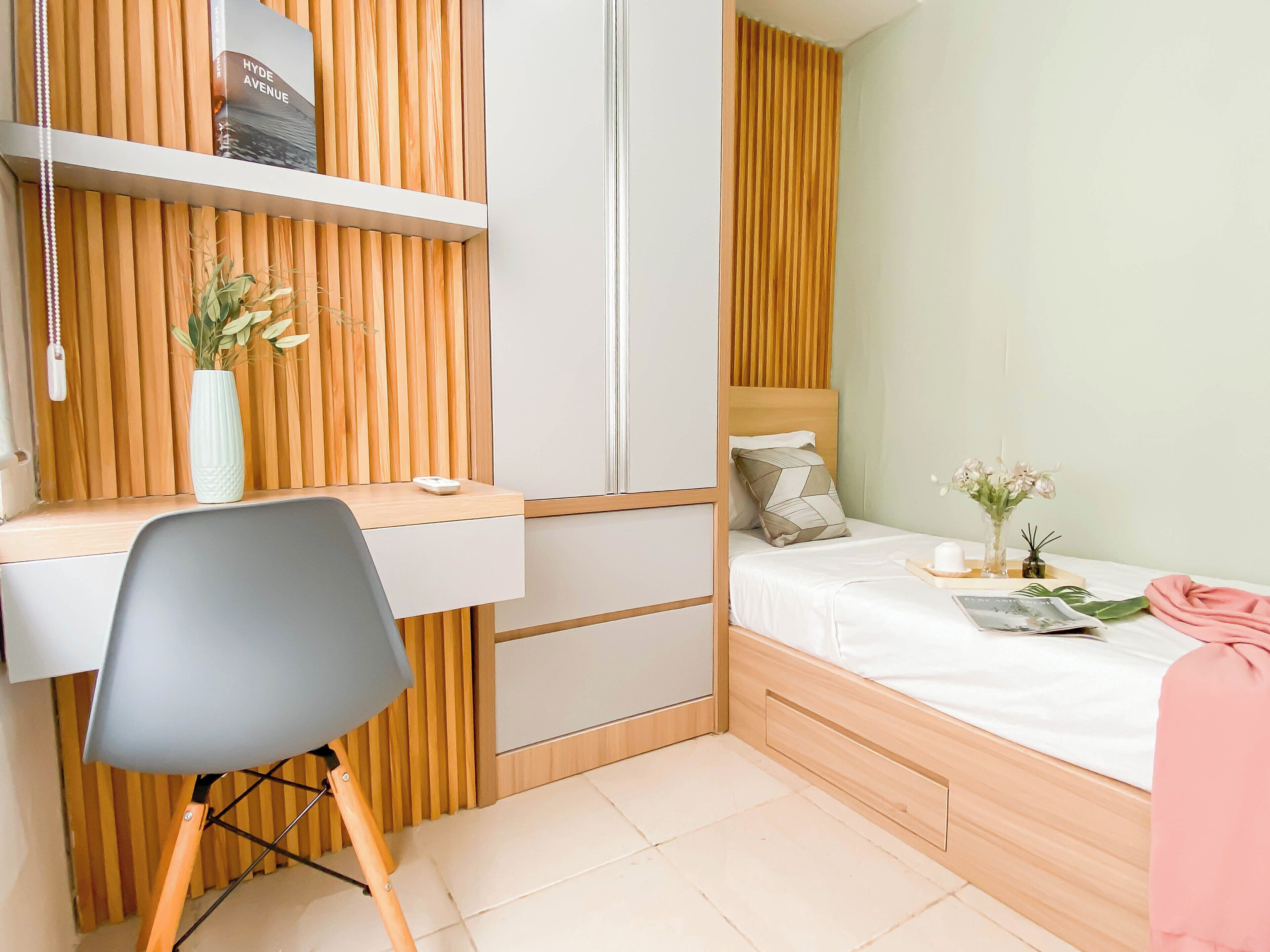 Quarto compacto com escrivaninha de madeira, uma cadeira, cama com travesseiro e mesa de cabeceira com um vaso de flores.