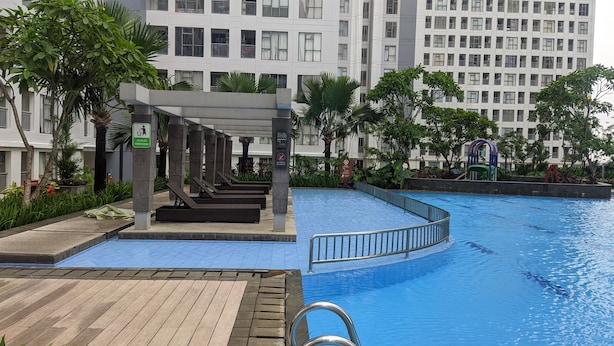 A swimming pool with a wooden deck area, surrounded by greenery and modern buildings. - Warm Studio Apartment M-Town Residence (Gading Serpong)