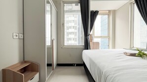 A bedroom with a bed, a window with curtains, a wooden bench, and a view of buildings outside. - Well Designed and Comfortable 3BR Nifarro Park Apartment (Jakarta)