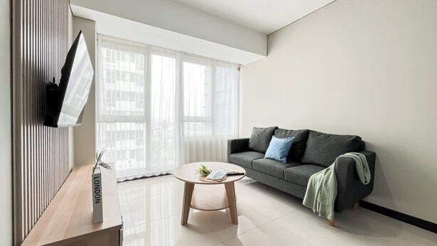 A modern living room with a grey sofa, a round wooden coffee table, and a TV mounted on the wall. - Well Designed and Comfortable 3BR Nifarro Park Apartment (Jakarta)