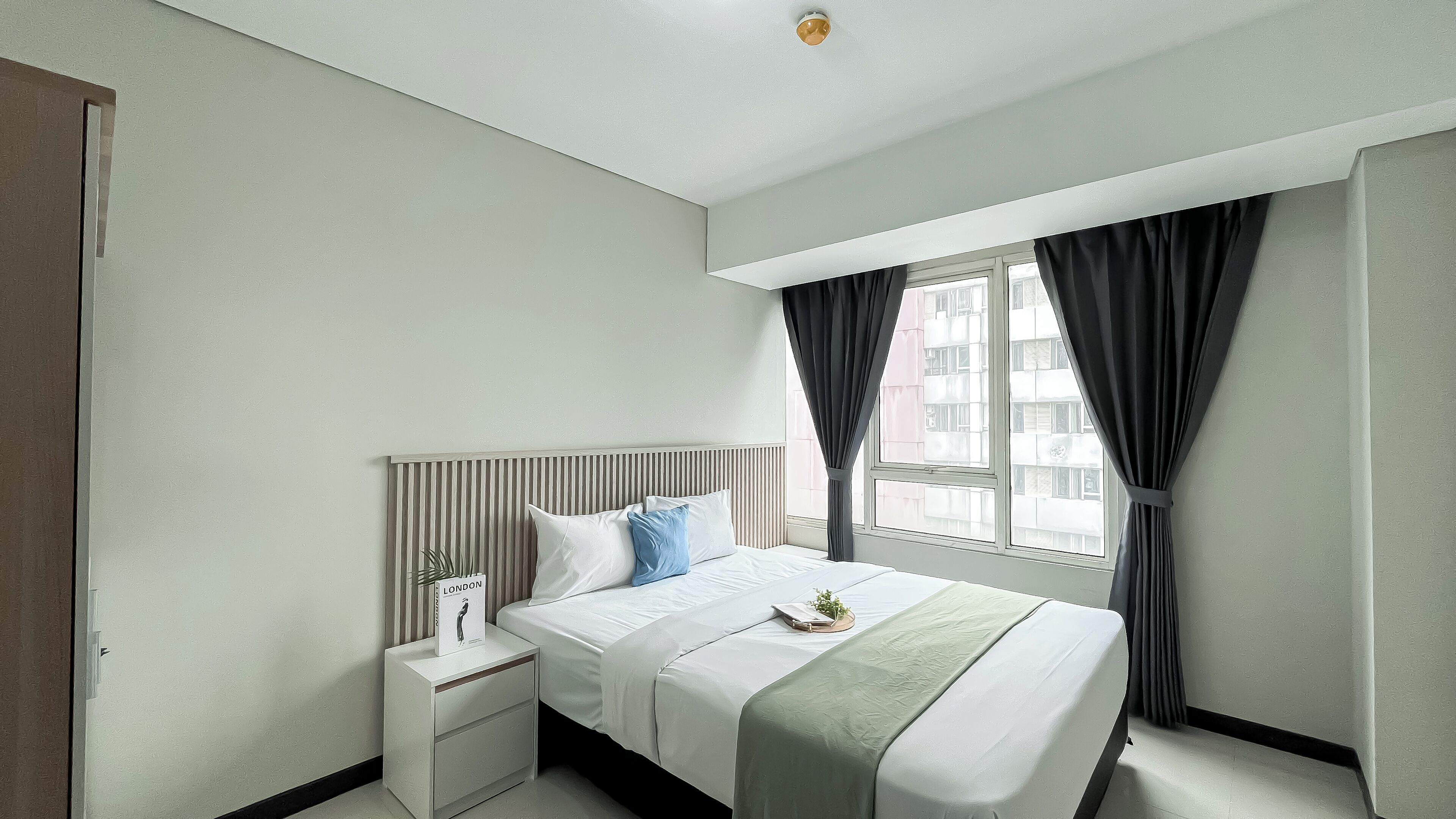 A hotel room with a large bed, a bedside table with a lamp, a window with curtains, and a view of buildings outside.