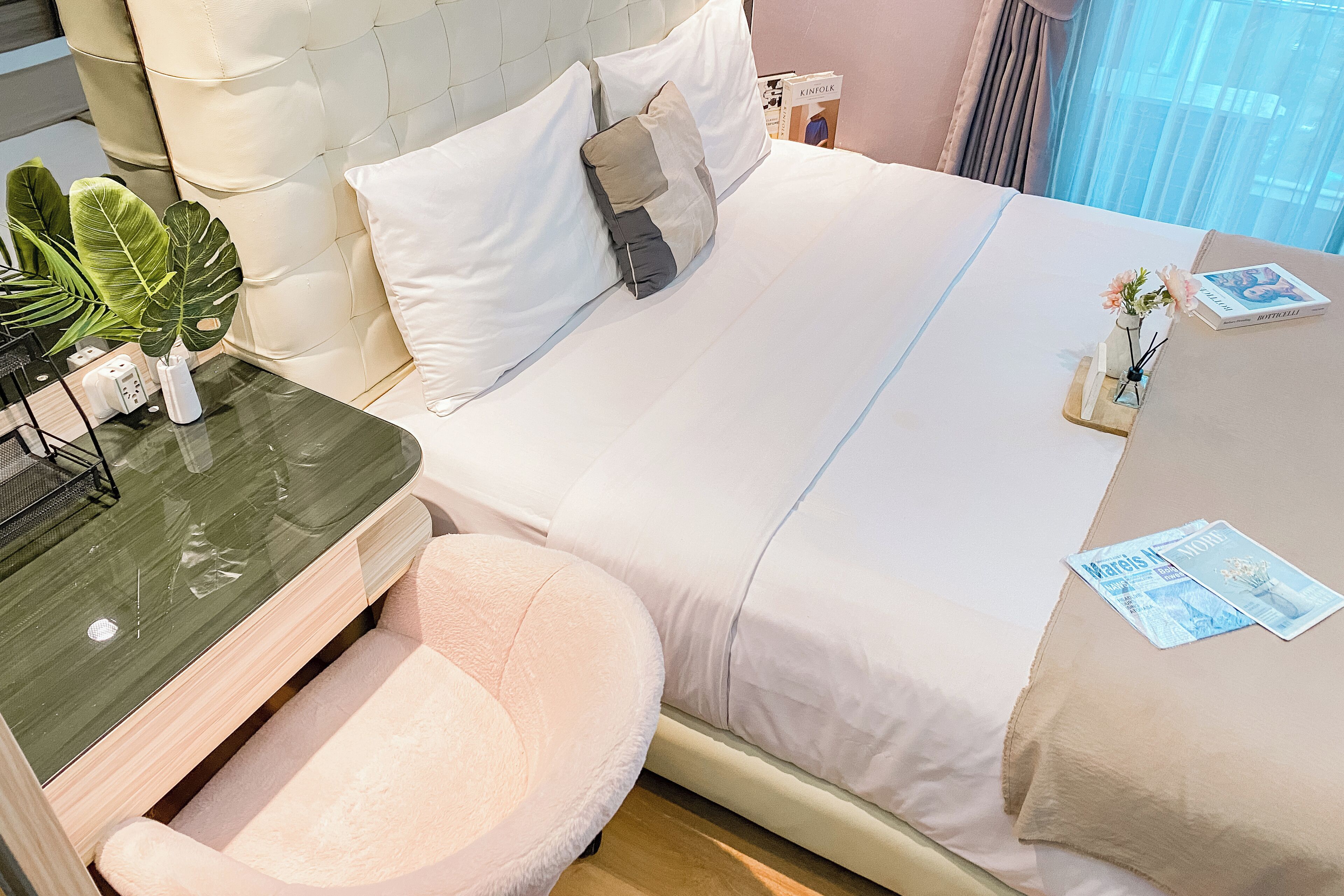 A neatly made bed with white bedding, a bedside table with a vase of flowers, and a small round chair.