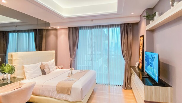 A modern hotel room with a large bed, a desk, a chair, a TV, and a balcony with curtains. - Spacious Studio Apartment at Capitol Suites (Jakarta)