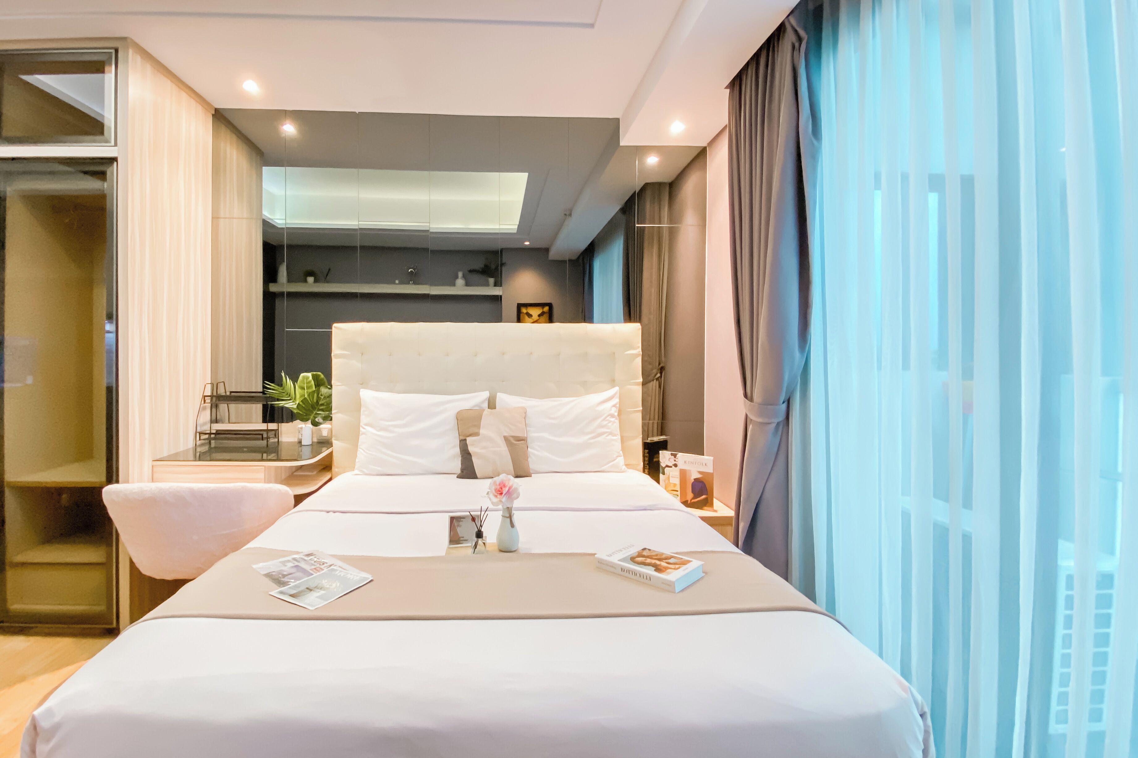 A modern hotel room with a large bed, bedside table, and a view of the city through sheer curtains.