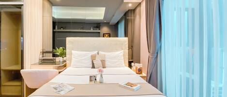 A modern hotel room with a large bed, bedside table, and a view of the city through sheer curtains.