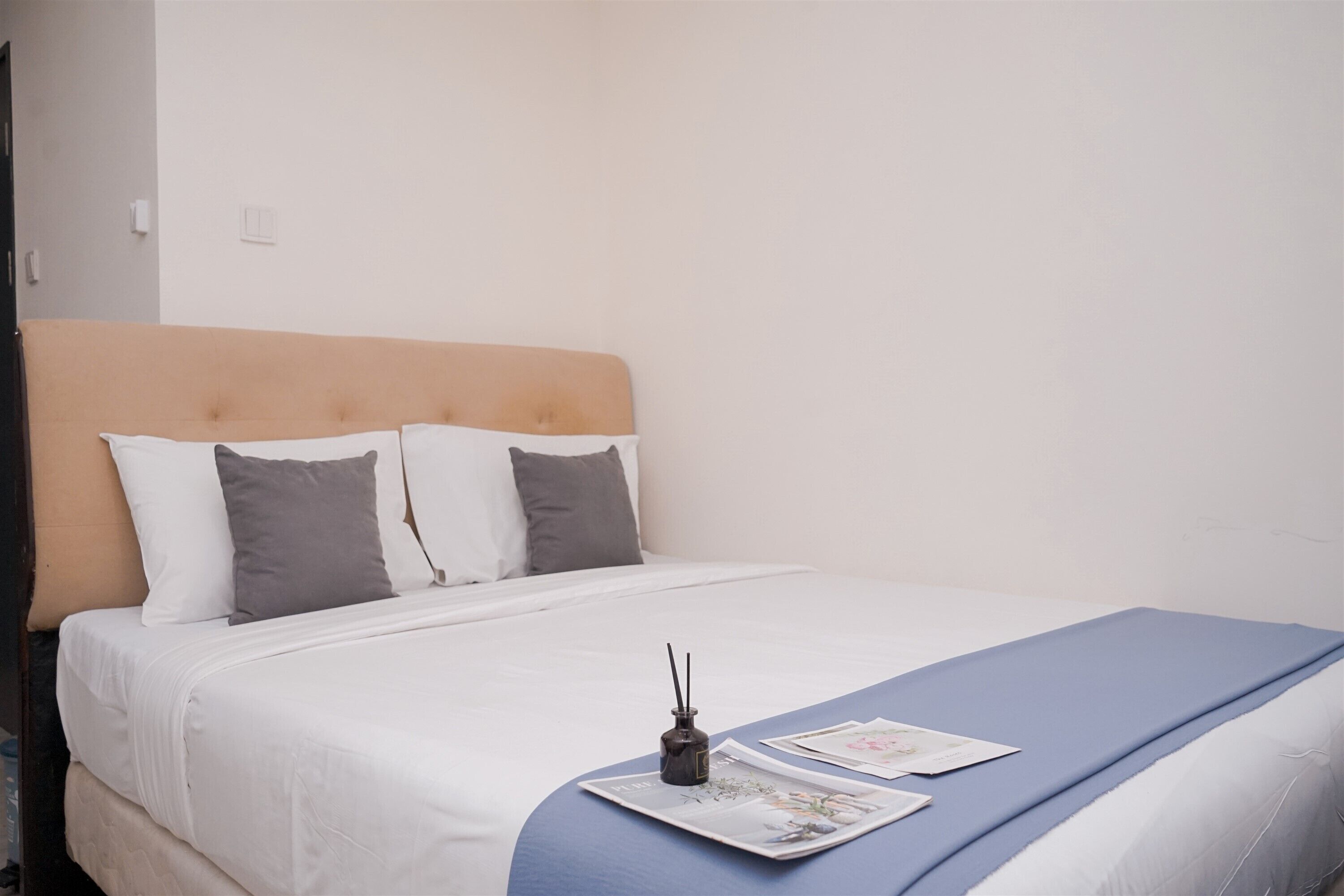 A neatly made bed with a blue bedspread and white linens, a small table with a book and a decorative item, and a wall with a light switch.