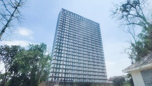 A tall, modern building with a glass facade, surrounded by greenery and a paved area in front. - Cozy Studio Apartment at Sudimara Forestwalk (South Tangerang)