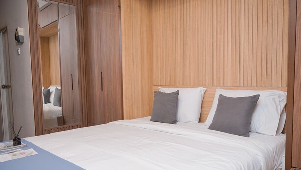 A bedroom with a wooden headboard, a bed with white bedding and gray pillows, a bedside table with a lamp, and a mirror on the wall. - Cozy Studio Apartment at Sudimara Forestwalk (South Tangerang)