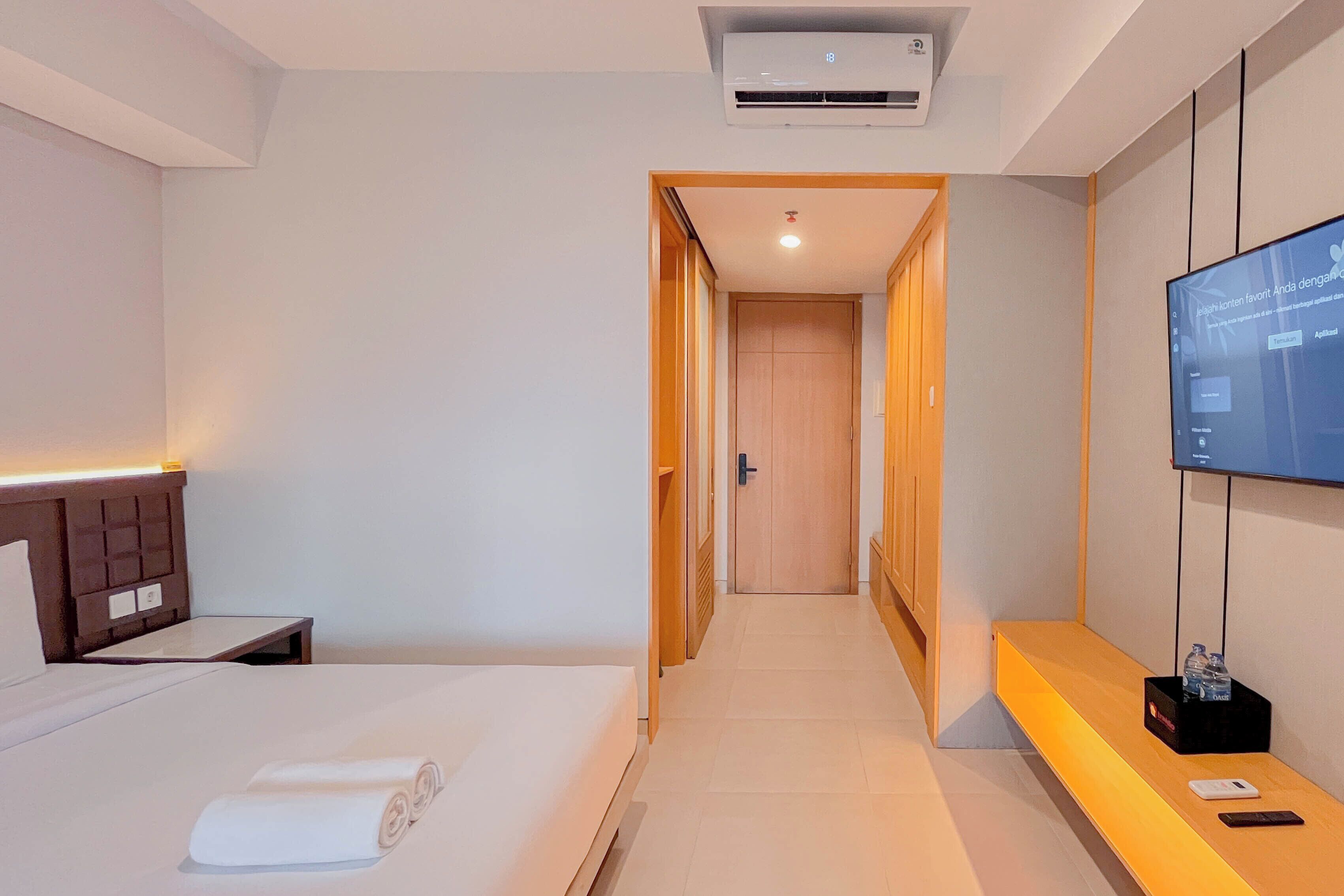Quarto de hotel com cama, mesa de cabeceira, televisão fixada na parede e um banco de madeira com uma garrafa de água e um telefone.
