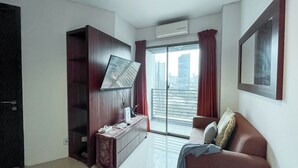 Una habitación de hotel moderna con televisor de pantalla plana en la pared, un armario de madera, una mesita con teléfono y control remoto, un sofá con cojines y un ventanal con vistas a la ciudad.