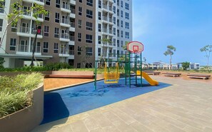 A playground with a basketball hoop and slides, surrounded by a landscaped area with benches and a building in the background. - Cozy and Good Place Studio Apartment Tokyo Riverside PIK 2 (Teluknaga)