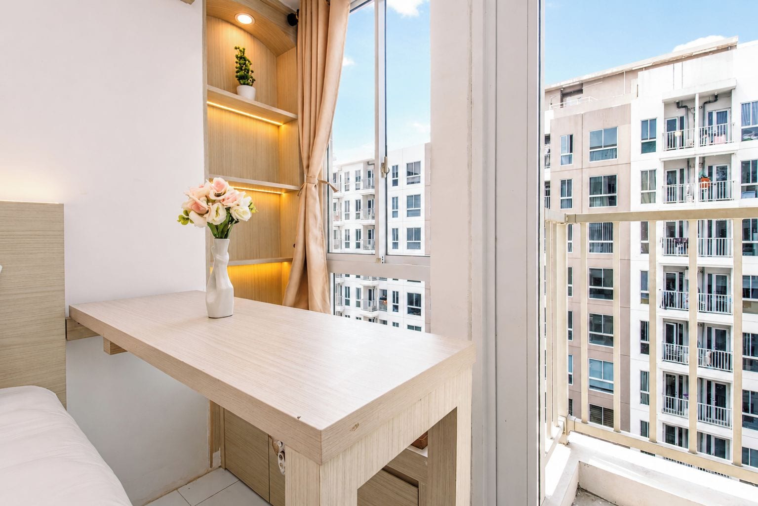 A modern bedroom with a wooden desk, a vase with flowers, and a view of a residential building.