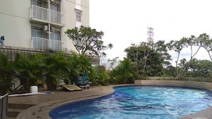 A swimming pool with a curved edge, surrounded by greenery and a building with balconies. - Nice and Cozy Studio at Bogor Valley Apartment (Bogor)