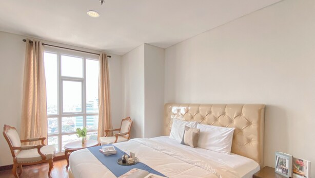 A hotel room with a large bed, a chair, a small table with a plant, and a window with curtains. - Big and Cozy 3BR Apartment Lucky Tower Residence (Jakarta)