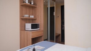 A hotel room with a bed, a microwave on a wooden shelf, and a door with a mirror. - Comfy and Minimalist Studio (No Kitchen) Apartment Great Western (Tangerang)