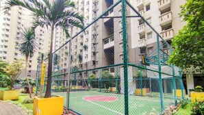 A fenced sports court with a basketball hoop, surrounded by greenery and tall residential buildings.