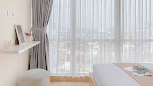 Ein Hotelzimmer mit einem Bett, einem kleinen Hocker, einem Regal mit Bilderrahmen und Blumen sowie Blick auf die Stadt durch transparente Vorhänge.
