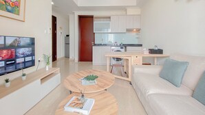 Ein modernes Wohnzimmer mit Flachbildfernseher, Couchtisch aus Holz und einer Couch mit Kissen. Im Hintergrund ist eine Küche mit hellen Schränken und einer Mikrowelle erkennbar.