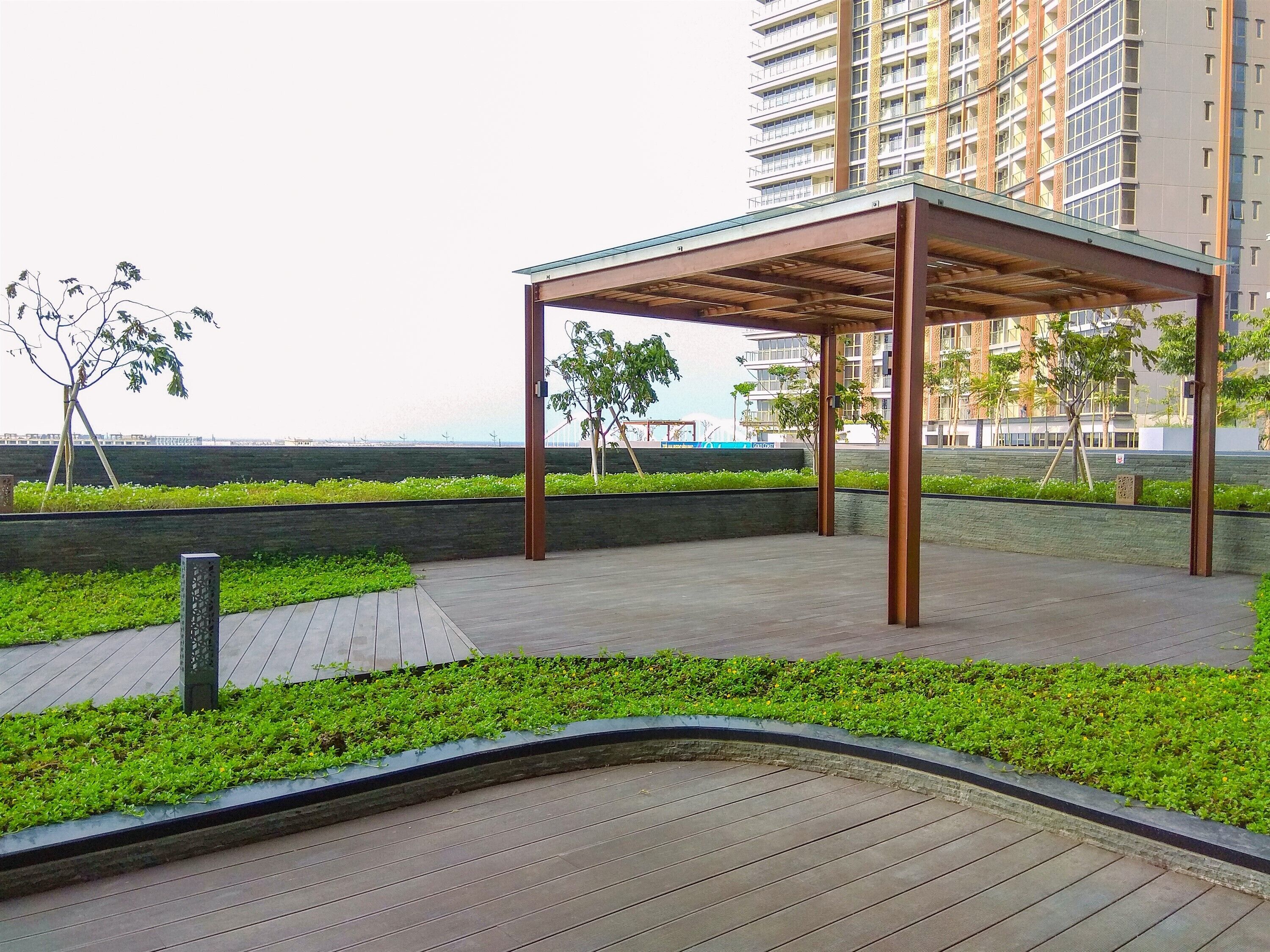 A covered outdoor area with a wooden structure, surrounded by greenery and a paved walkway.