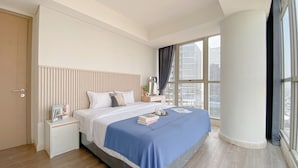 A modern hotel room with a large bed, a nightstand, a bedside table, and a view of the city through large windows. - Fully Furnished and Elegant 2BR at Gold Coast Apartment PIK (Jakarta)