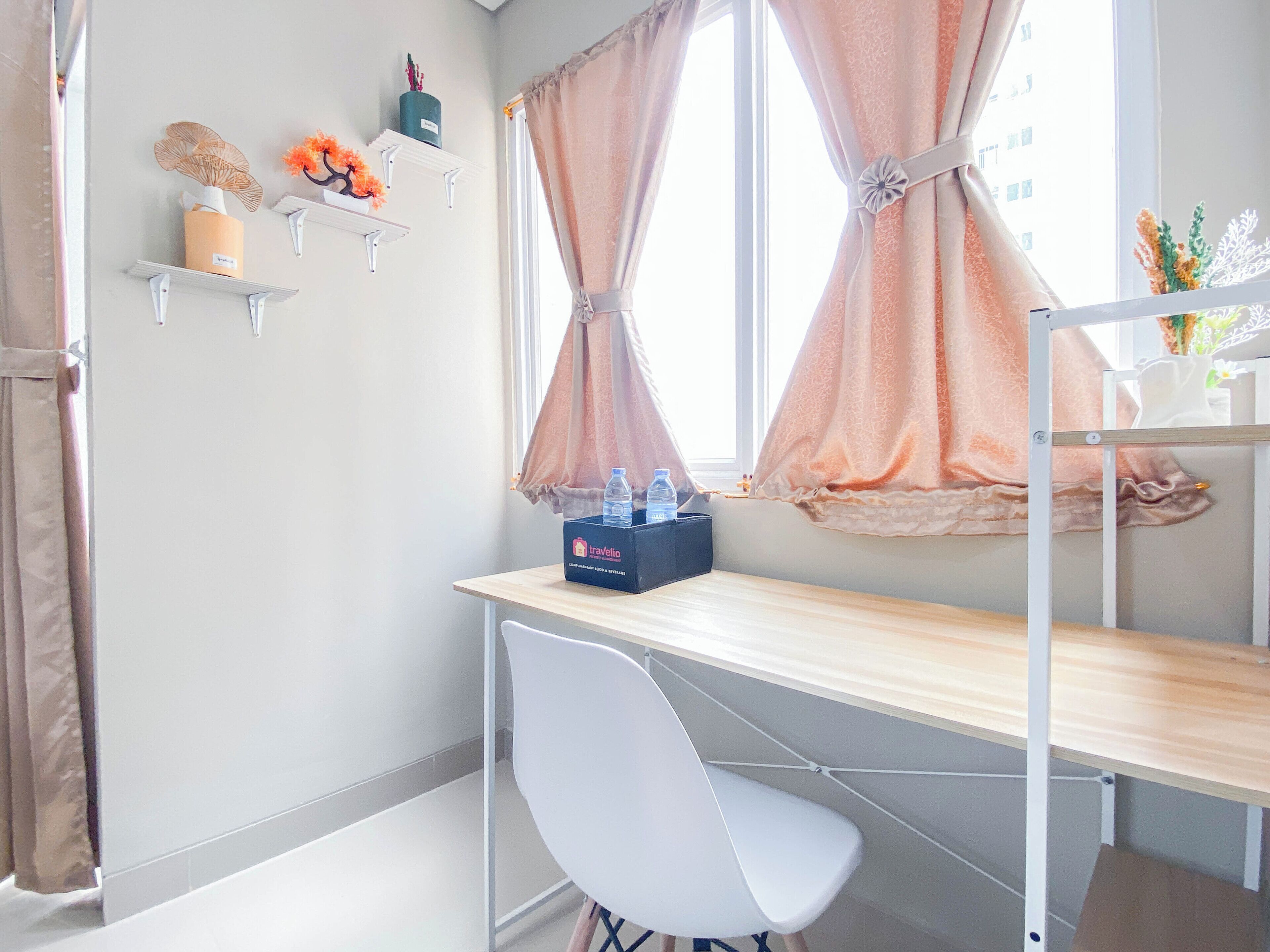 A room with a wooden desk, a white chair, a water bottle, and a box labeled 'Travelio'. Shelves with decorative items are on the wall.