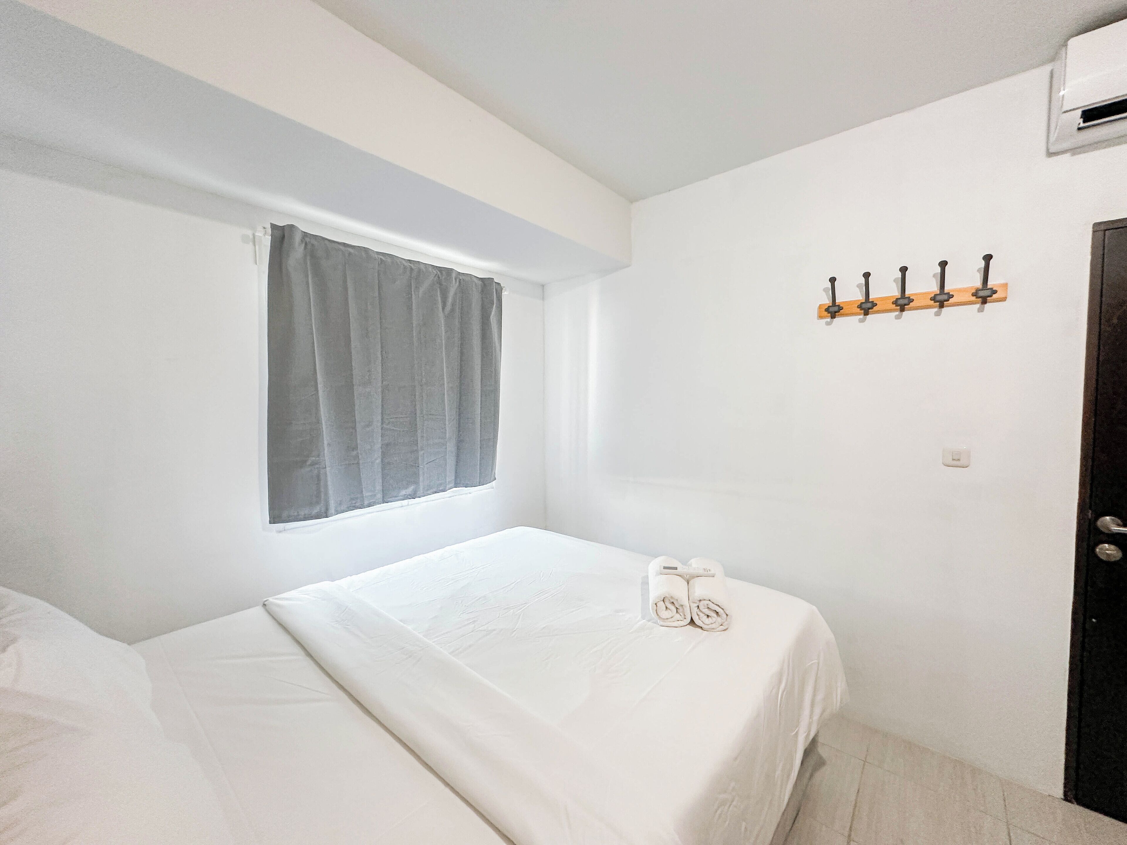A small, clean room with a single bed, a window with a grey curtain, a wall-mounted towel rack, and a door.