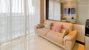 Un salon moderne avec un canapé beige, de grandes fenêtres ornées de rideaux légers, un téléviseur à écran plat fixé au mur et un meuble multimédia noir au design épuré.