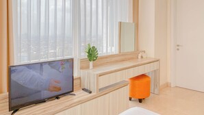 Une chambre moderne équipée d’une télévision, d’un comptoir en bois, d’un miroir et d’une grande fenêtre agrémentée de rideaux légers.