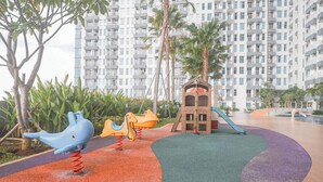 A playground with a slide and swings, surrounded by palm trees and a multi-story building in the background. - Cozy and Spacious 1BR at Anderson Supermall Mansion Apartment (Surabaya)