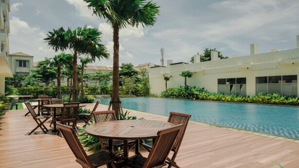 A poolside area with wooden tables and chairs, surrounded by palm trees and a swimming pool.