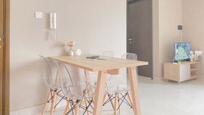 A modern dining area with a wooden table, clear chairs, a small vase with flowers, a wall-mounted phone, and a TV on a wooden stand. - Good Deal and New Furnished 2BR Ciputra International Apartment (Jakarta)