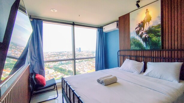 A modern hotel room with a large bed, a view of the city, and a painting on the wall. - Wonderful Thematic 2BR at Praxis Apartment (Surabaya)