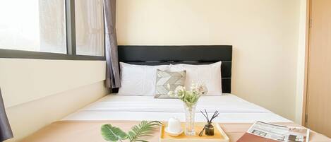 A neatly made bed with white bedding, a black headboard, and a bedside table with a tray holding a vase of flowers, a candle, and a book.