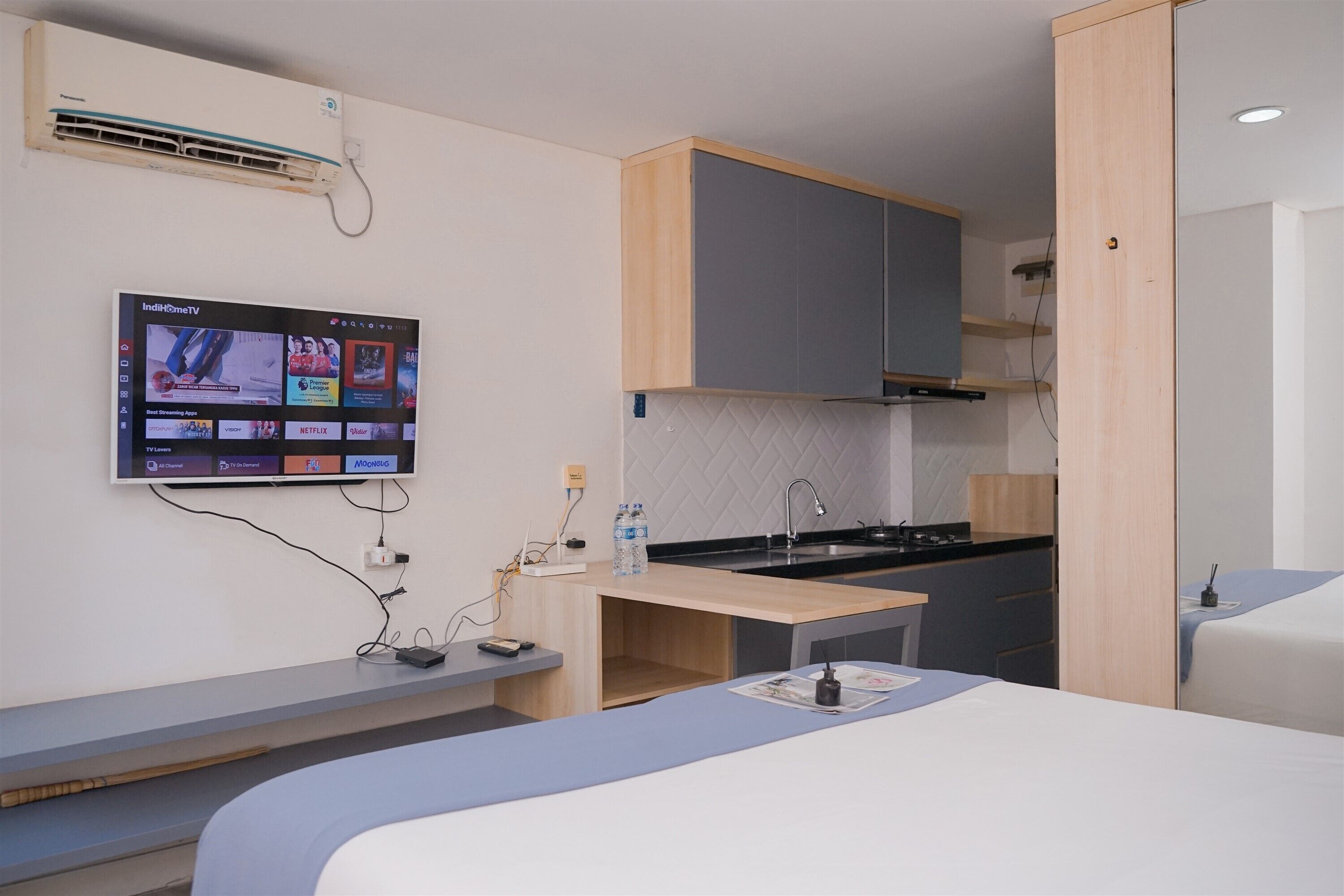 A compact hotel room with a bed, a small desk, a kitchenette, and a mounted TV displaying streaming services.