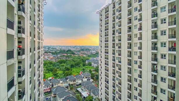 A high-rise residential building with multiple balconies, a view of a residential area with houses, and a sunset in the background. - Good Studio Room at Cinere Resort Apartment (Depok)