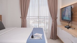 Una habitación de hotel con una cama, un televisor sobre un mueble de madera y una ventana con cortinas translúcidas.