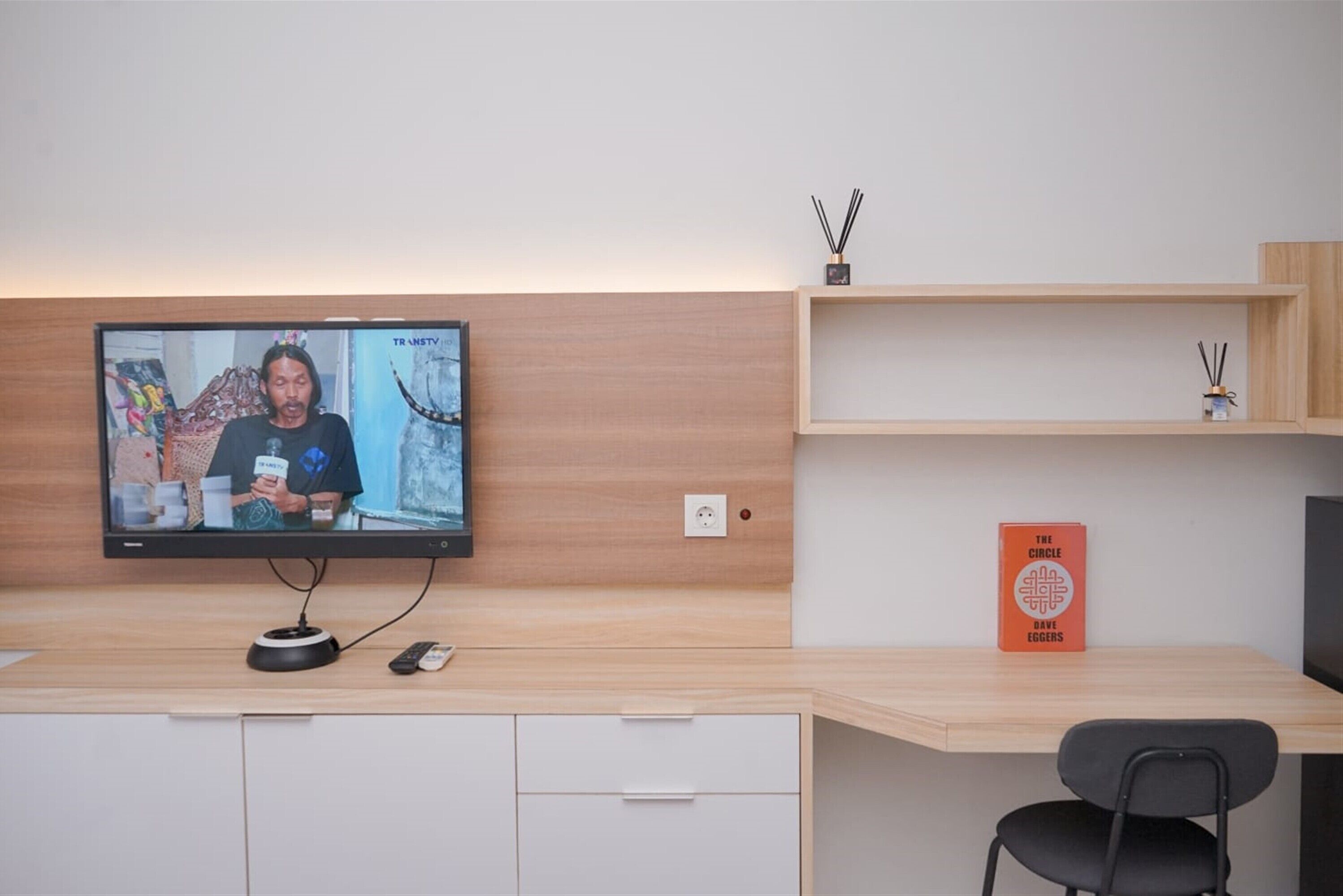 Ein modernes Zimmer mit einem an einer Holzwand montierten Flachbildfernseher, einem Schreibtisch mit Stuhl und einem Regal mit einem Buch mit dem Titel 'The Circle' von Dave Eggers.