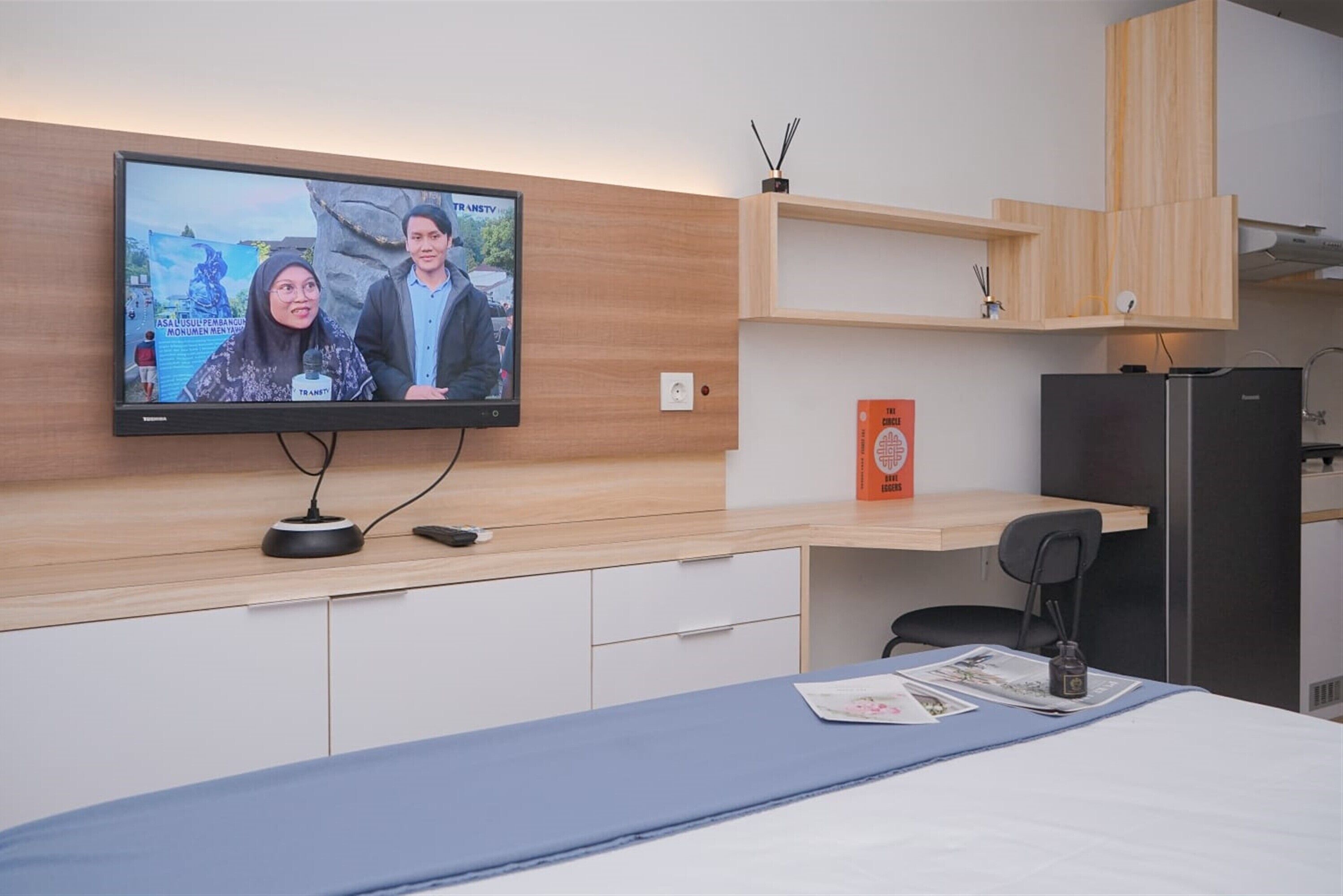 A modern hotel room with a flat-screen TV mounted on the wall, a wooden desk, a chair, a bed with a blue bedspread, and a small shelf with a box and a bottle.