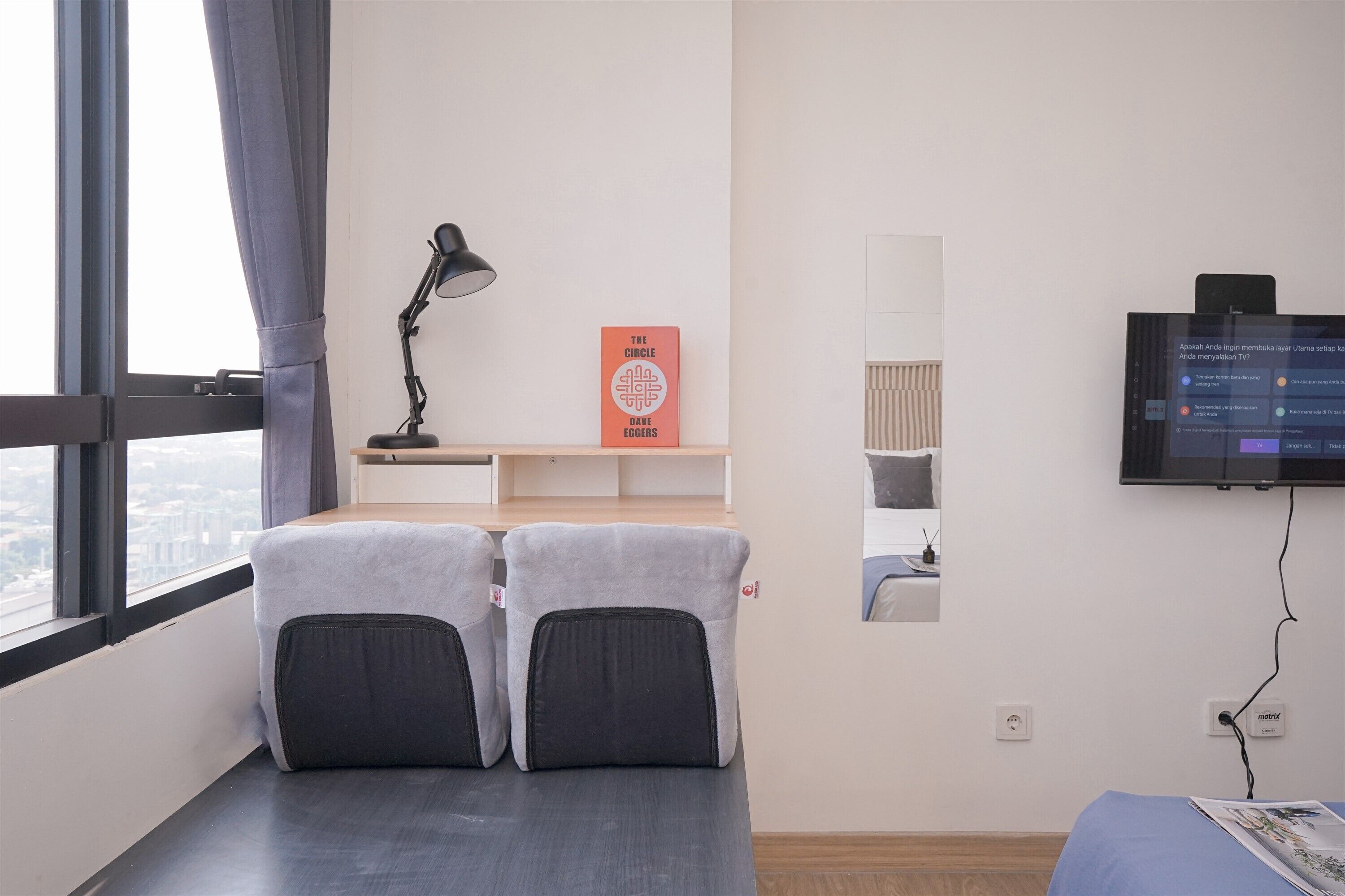 Quarto de hotel com uma cama, uma mesa de trabalho com abajur, uma televisão e uma janela com cortinas.