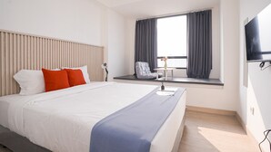 Quarto de hotel moderno com uma cama grande, uma mesinha de cabeceira, uma cadeira, uma janela com cortinas e uma TV de tela plana fixada na parede.
