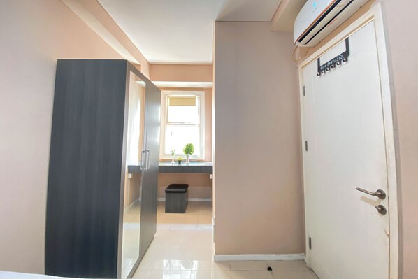 A hallway with a door, a trash bin, and a window with plants. - Pleasurable 2BR Apartment Parahyangan Residence (Bandung)
