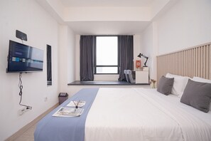 A modern hotel room with a large bed, a TV mounted on the wall, a window with curtains, and a bedside table with a lamp. - Warm Cozy Studio Room at Collins Boulevard Apartment (Tangerang)