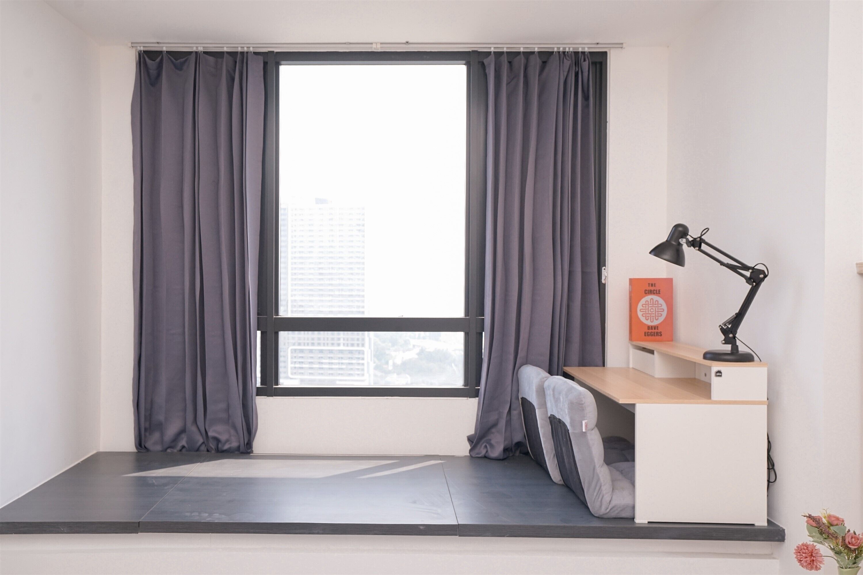 A room with a large window, grey curtains, a desk with a lamp, and a small shelf with a book.