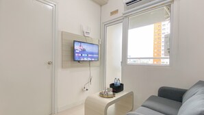 A modern living room with a grey sofa, a flat-screen TV mounted on the wall, a small white side table with a water bottle, and a window with a city view. - Good and Cozy 2BR at Green Pramuka City Apartment (Jakarta)