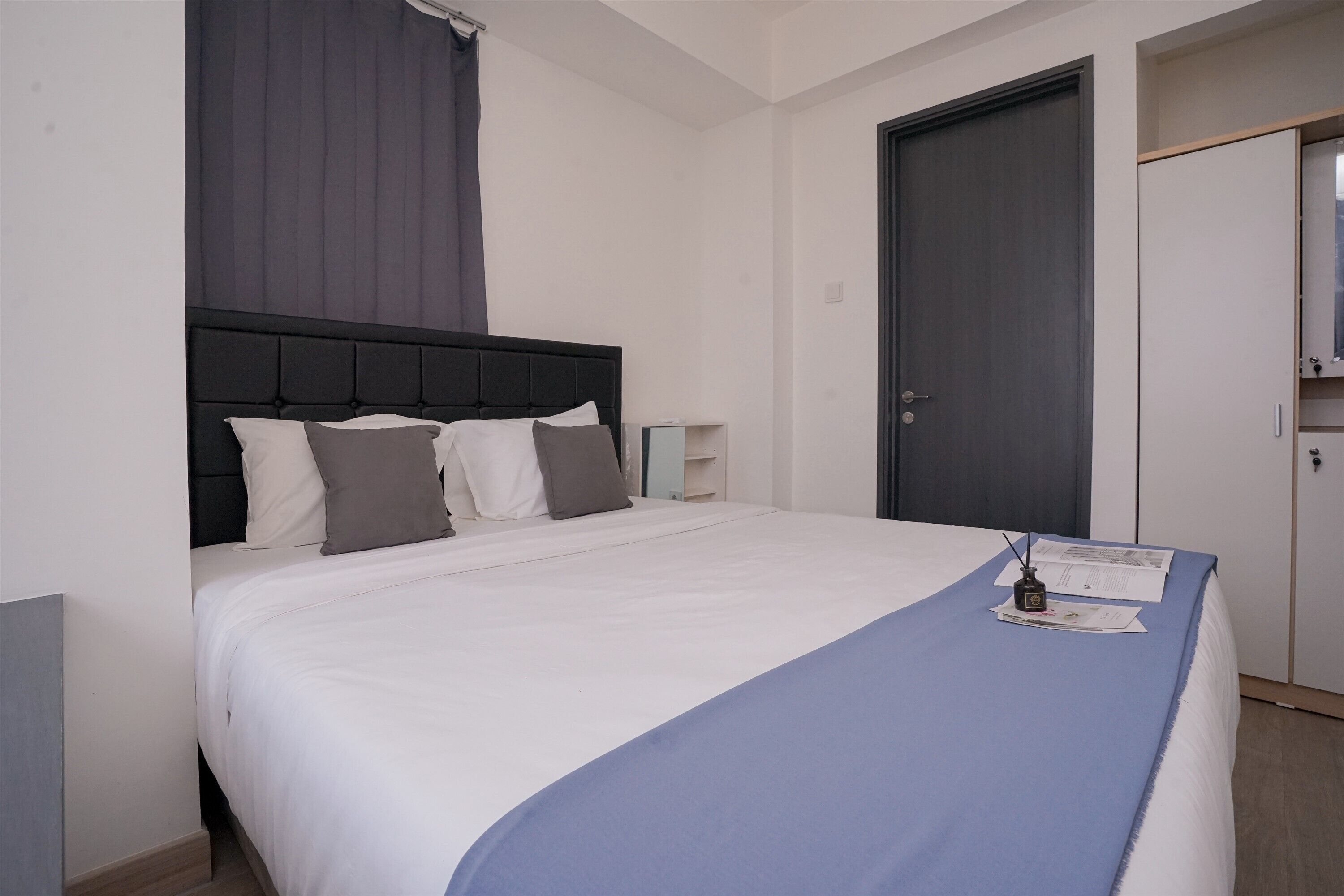 Una cama bien hecha con colcha azul y sábanas blancas, una mesita de noche con lámpara, y una puerta cerrada en una habitación de hotel.