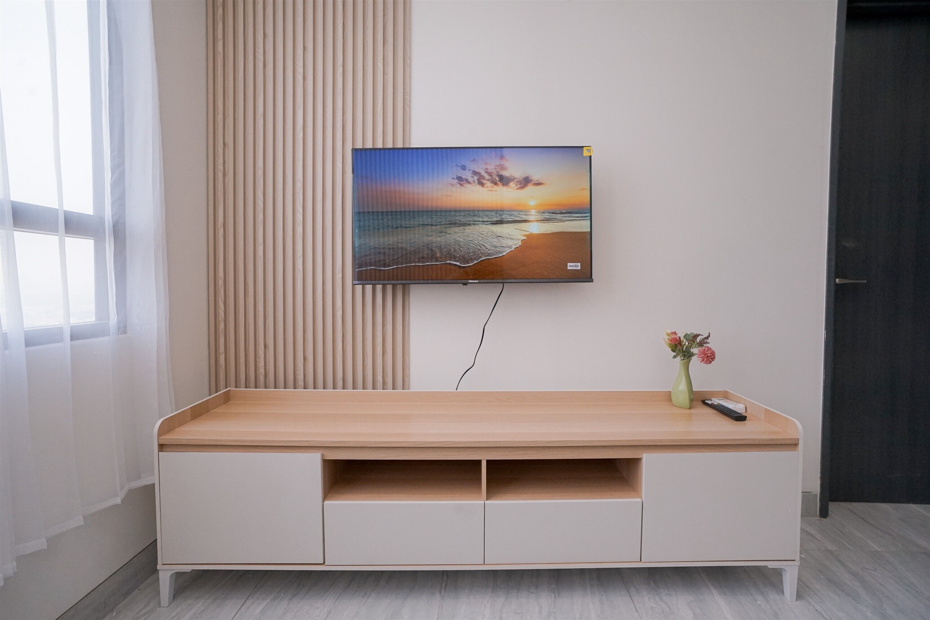 Una sala de estar moderna con un mueble de entretenimiento de madera, un televisor montado mostrando una puesta de sol en la playa y un jarrón con flores sobre el mueble.